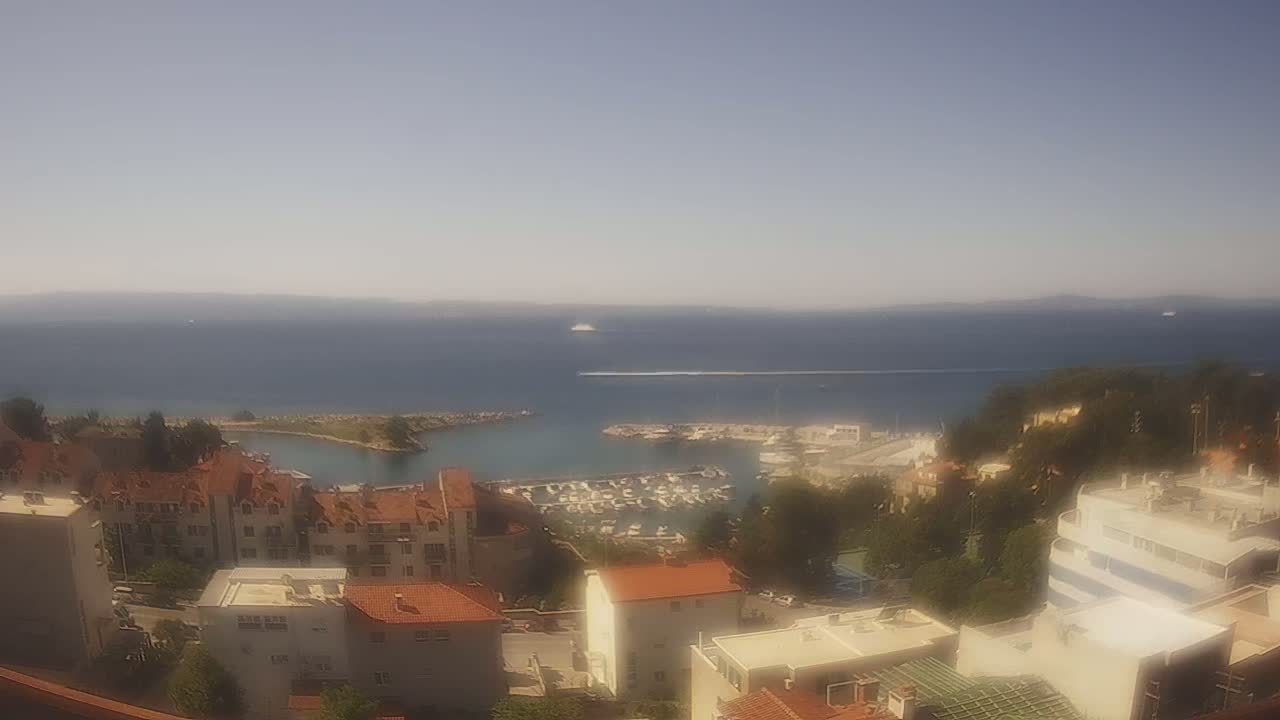 A hazy, sunny day overlooks a coastal town with red-roofed buildings, a marina filled with boats, and a calm sea extending to a distant horizon.
