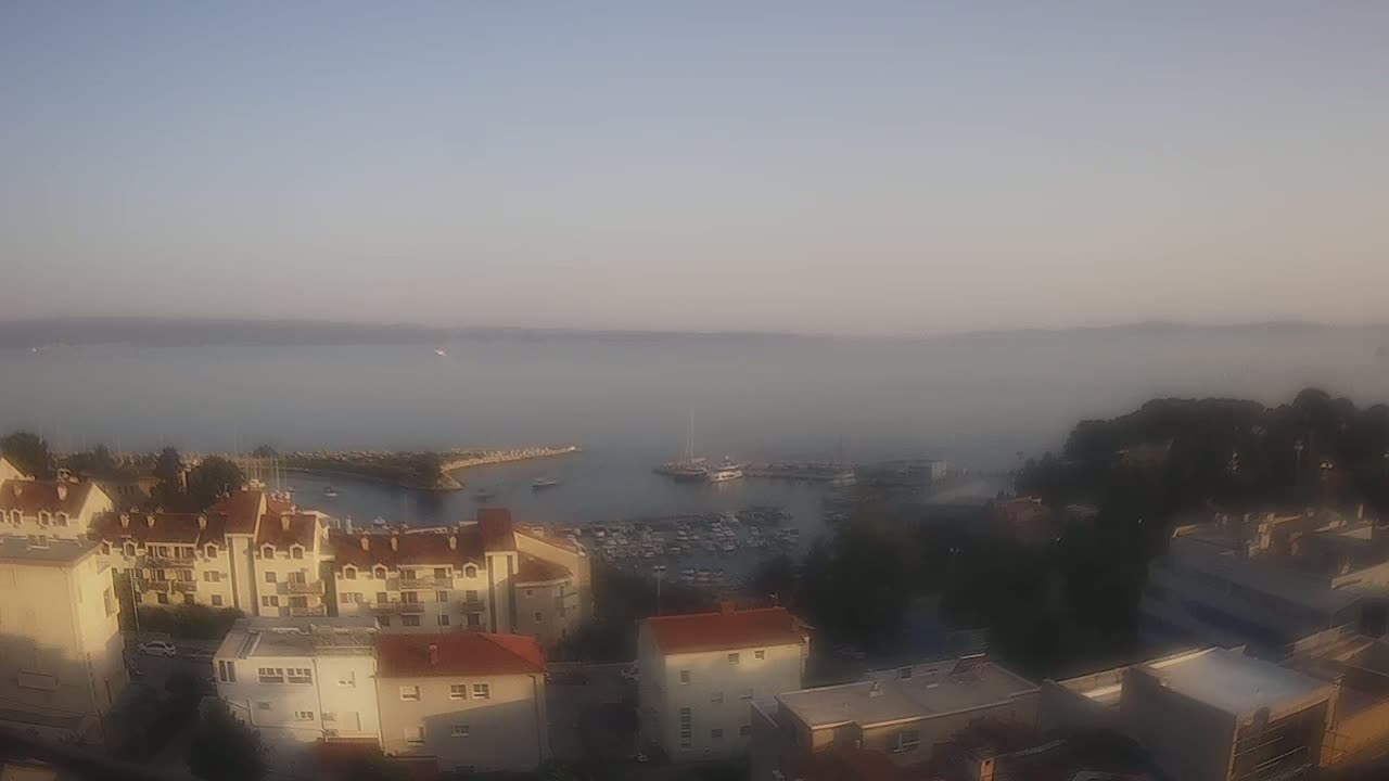 A hazy sunrise reveals a harbor filled with boats, bordered by buildings and a stretch of calm water.