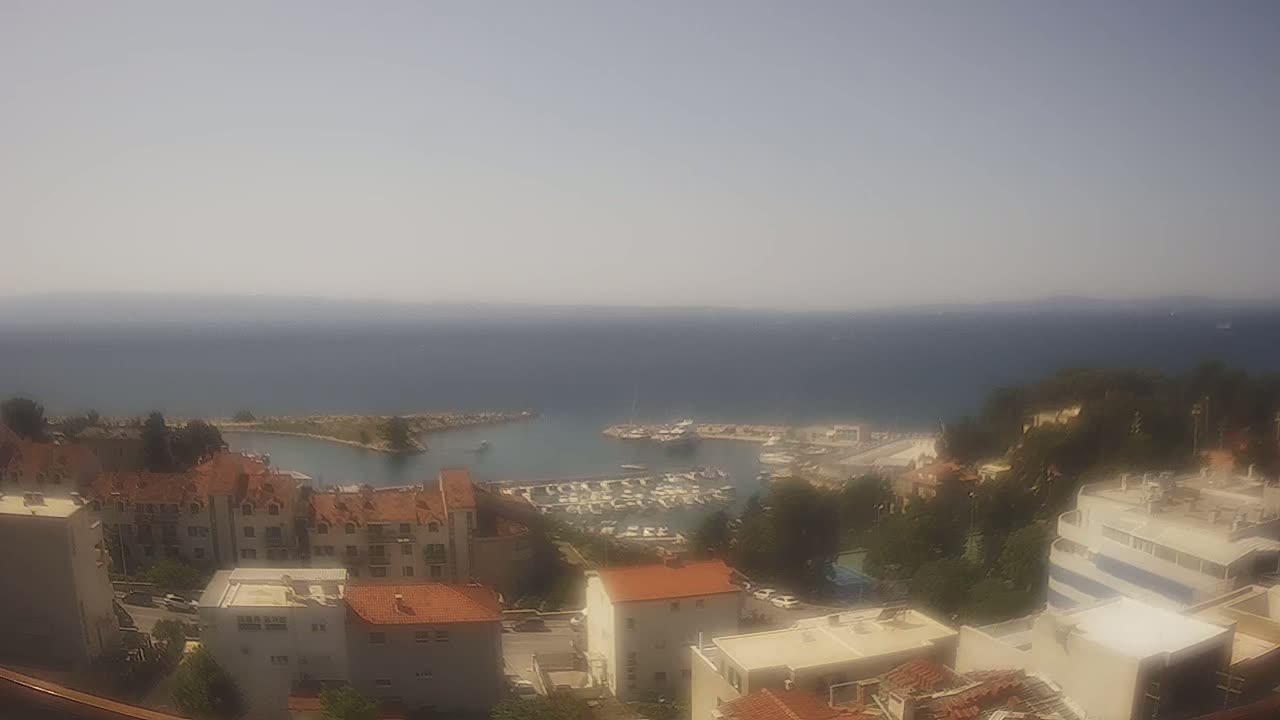 A hazy, sunny day overlooks a coastal town with a marina filled with boats, buildings with red tile roofs, and green trees, all situated next to a calm body of water.