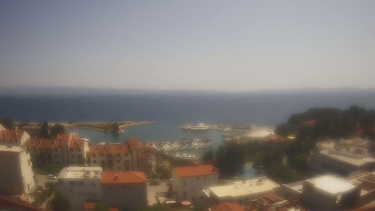 A hazy, sunny day overlooking a calm, blue sea with a marina full of boats and a collection of low-rise buildings nestled on a hillside.