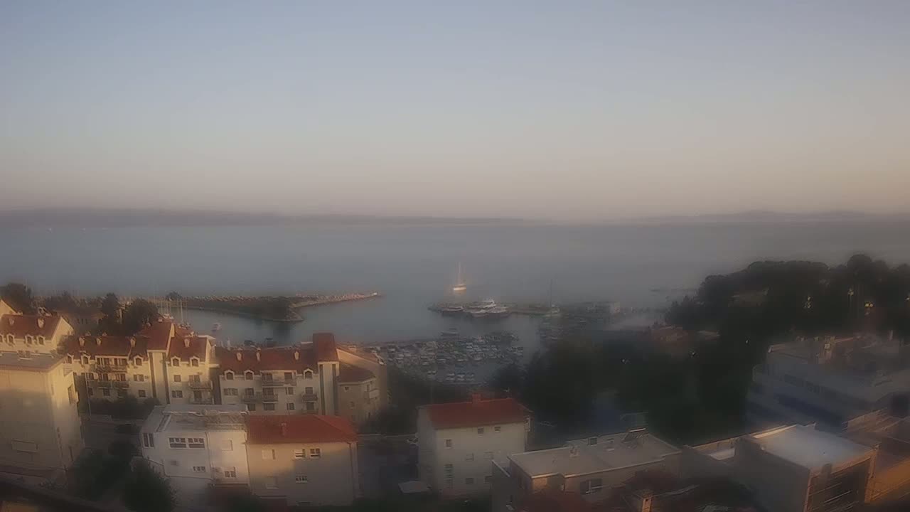 A hazy sunrise illuminates a coastal town overlooking a calm harbor filled with boats.