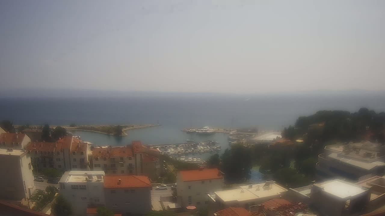 A hazy, sunny day overlooks a calm sea with a marina filled with boats and a small town of red-roofed buildings.