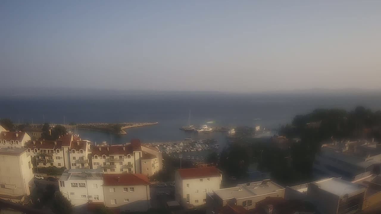 A hazy, sunny day overlooks a calm harbor filled with boats, nestled between buildings and green vegetation.