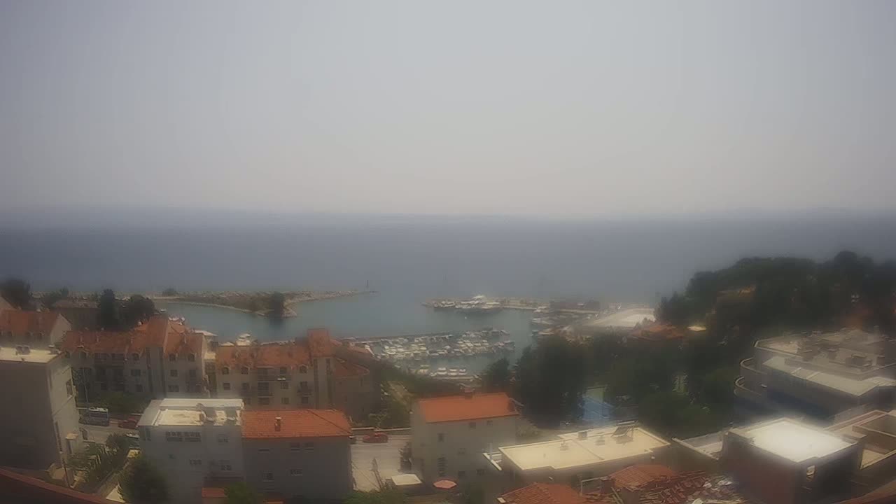 An aerial view of a coastal town on a hazy day shows a harbor filled with boats, and buildings with red-tiled roofs nestled amongst green trees.
