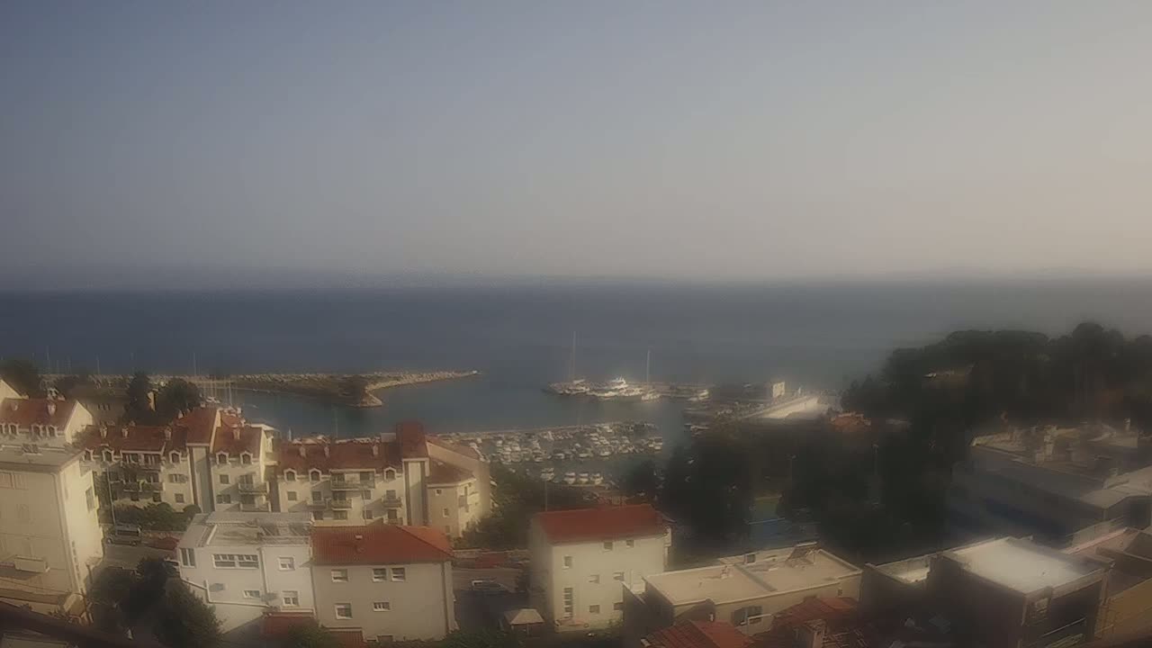A hazy, sunny day overlooks a coastal town with red-roofed buildings, a marina filled with boats, and a calm sea extending to the horizon.