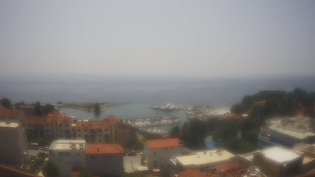A hazy, sunny day overlooks a coastal town with a marina filled with boats, and buildings nestled amongst green trees.