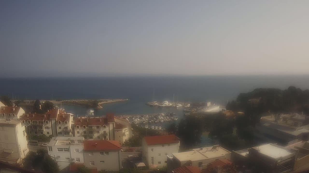A hazy, sunny day overlooks a coastal town with a harbor filled with boats, and buildings with red tile roofs nestled close to the water.