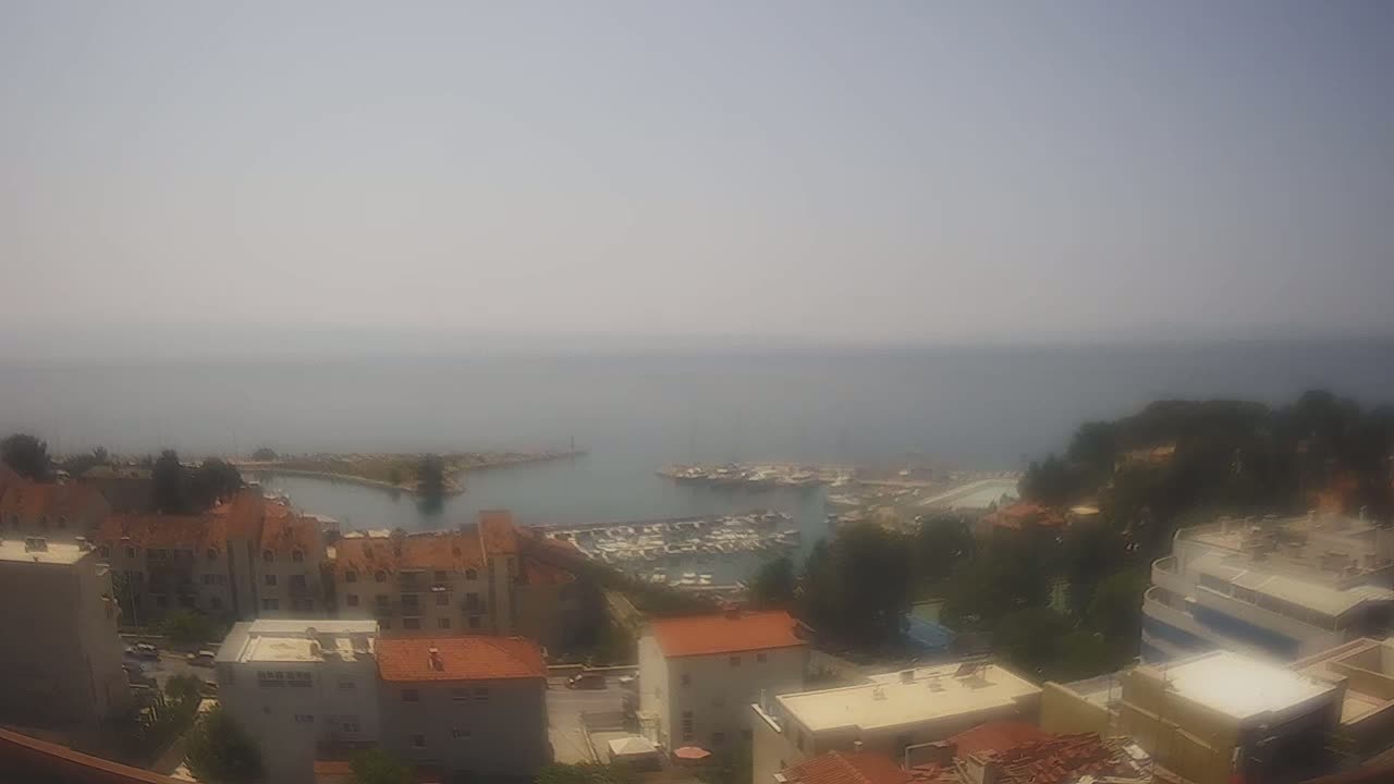 A hazy, sunny day overlooking a coastal town with a marina filled with boats, and buildings with red-tiled roofs.