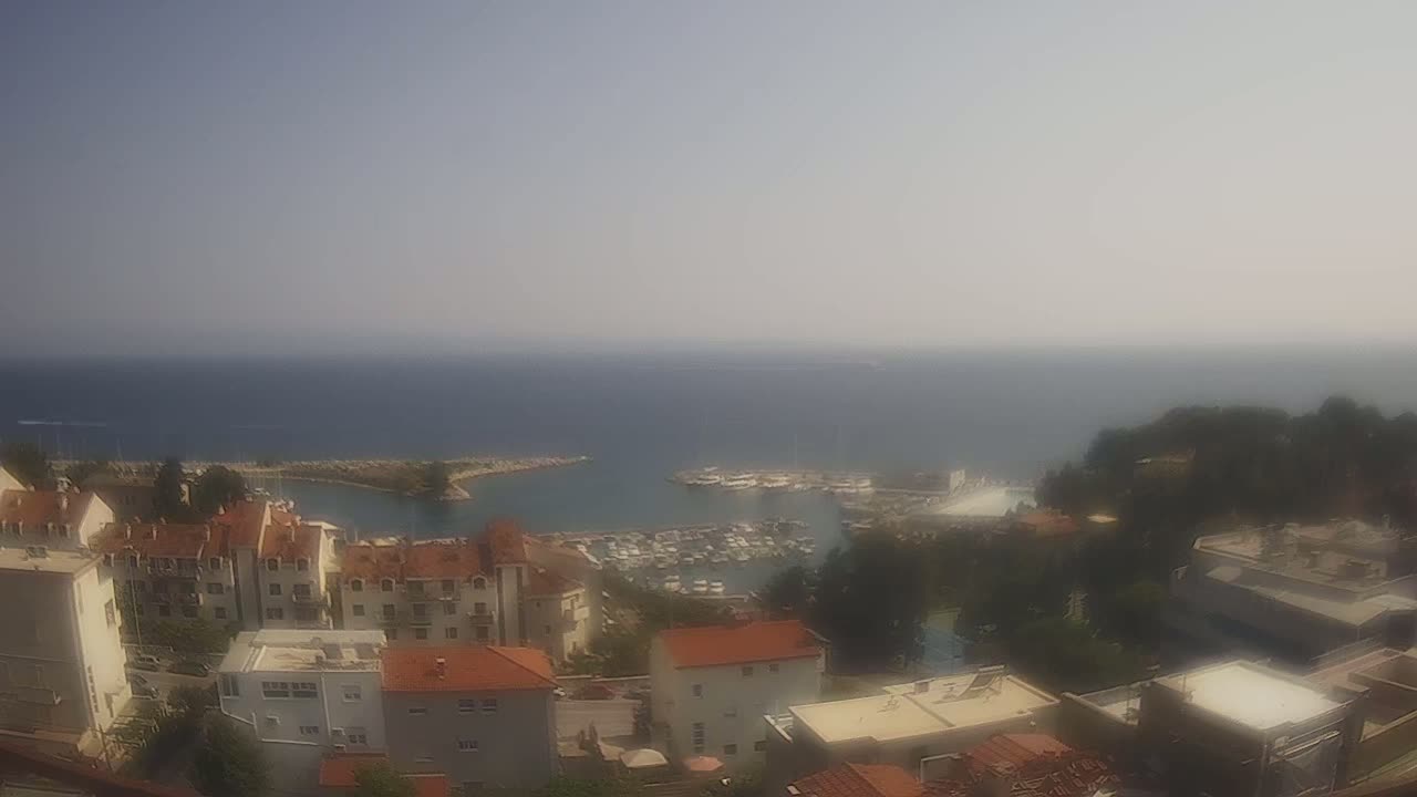 A hazy, sunny day overlooks a marina filled with boats, nestled between a hillside of buildings and a calm sea.