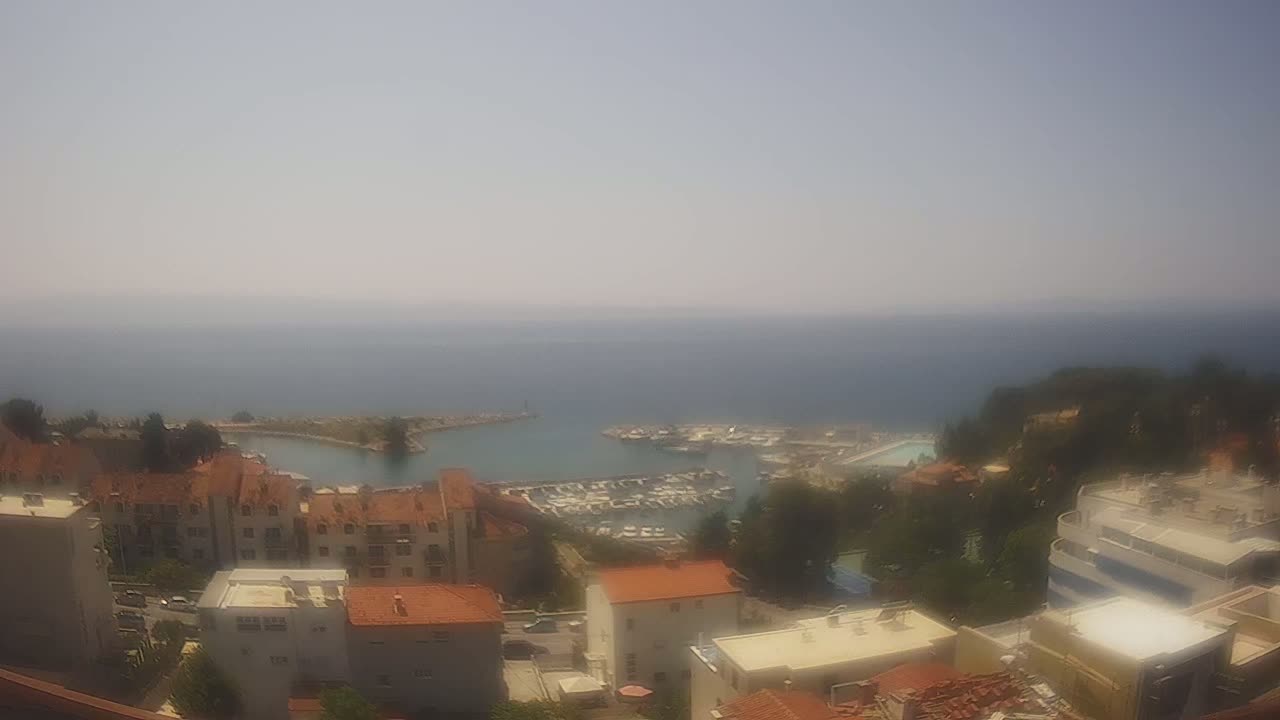 A hazy, sunny day overlooks a coastal town with a marina filled with boats, and buildings with red-tiled roofs.