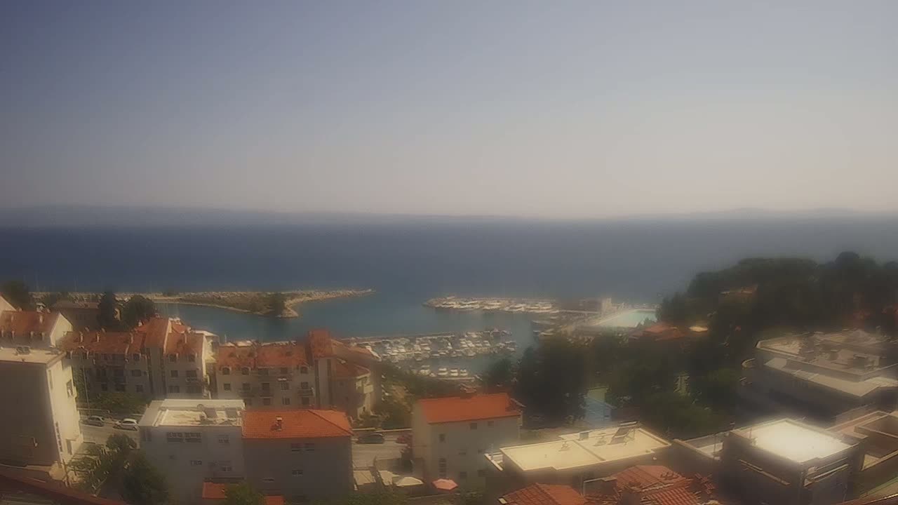 A hazy, sunny day overlooks a coastal town with a marina filled with boats, and red-roofed buildings cascading down a hillside towards the sea.