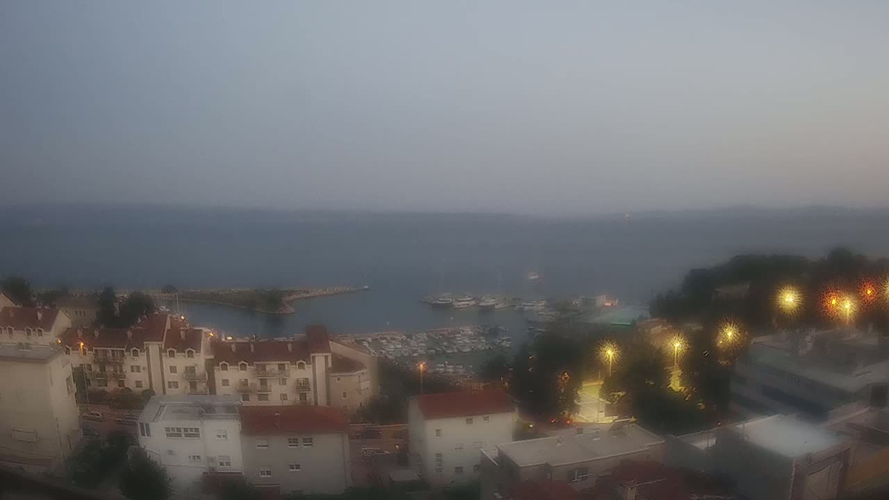 A hazy, sunny day overlooks a coastal town with a marina filled with boats, and red-roofed buildings cascading down a hillside towards the sea.