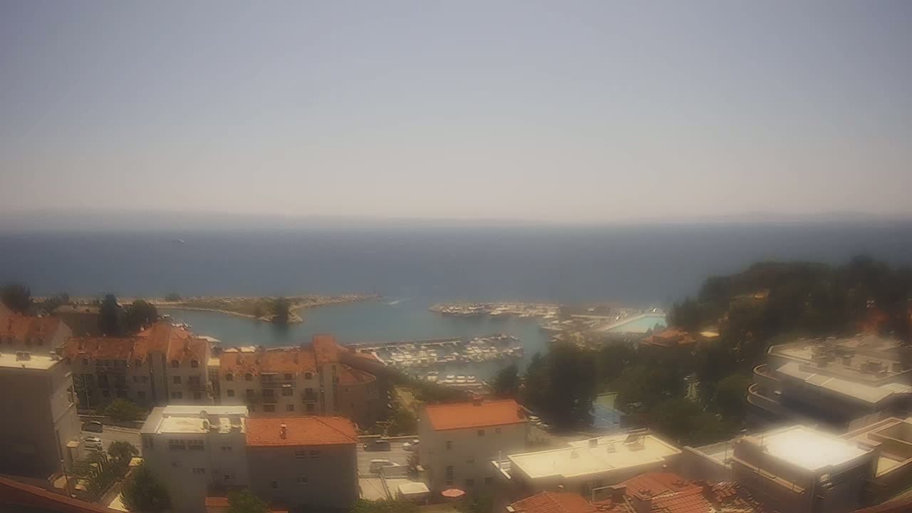 A hazy, sunny day overlooking a coastal town with a marina filled with boats, and numerous buildings with red tile roofs.
