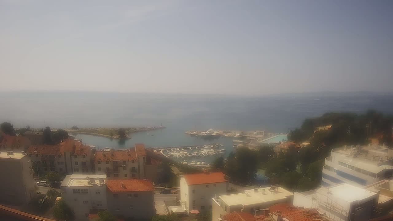 A hazy, sunny day overlooks a calm sea harbor filled with boats and surrounded by low-rise buildings.