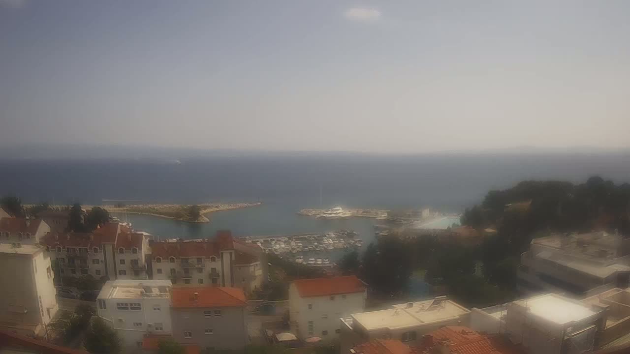 A hazy, sunny day reveals a coastal town with a marina filled with boats, nestled amongst buildings on a hillside overlooking the calm sea.