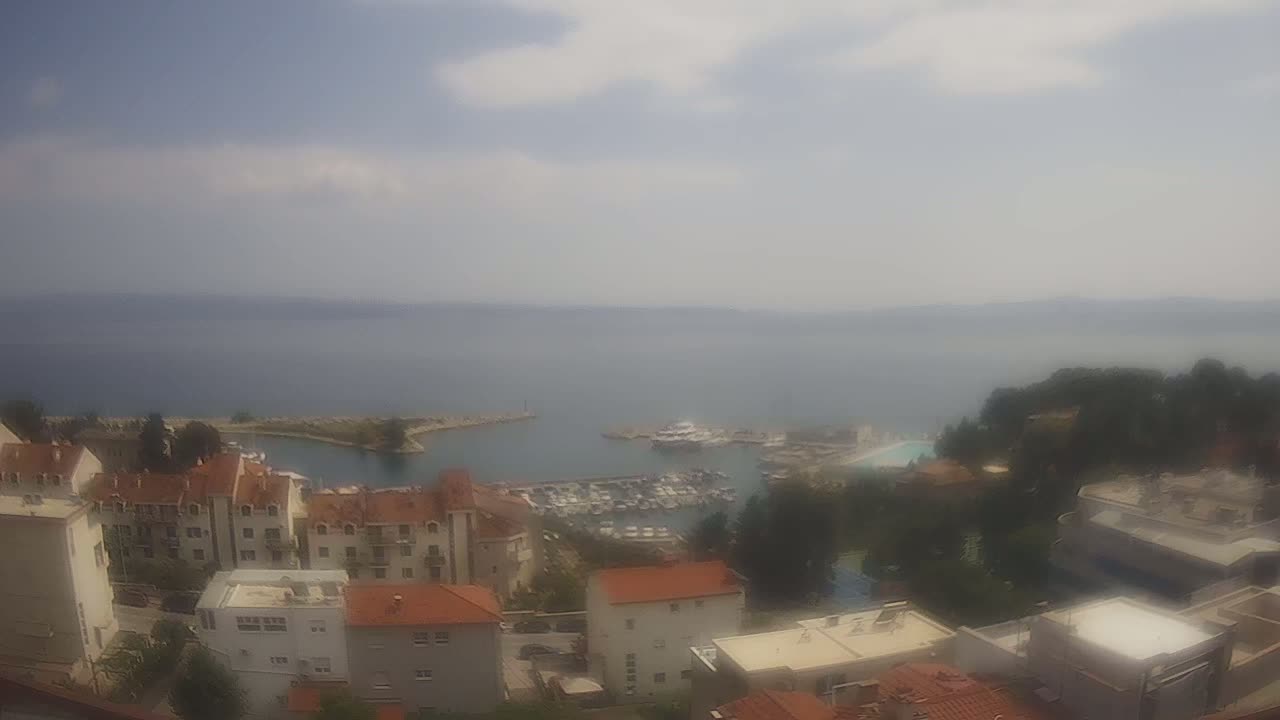 A hazy, partly cloudy day overlooks a marina filled with boats, nestled between a hillside town of low-rise buildings with red tile roofs and a calm, expansive sea.