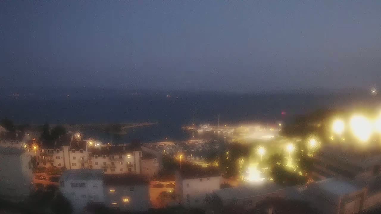 A nighttime view of a coastal town with a marina, illuminated buildings, and a calm, dark sea under a dusky sky.