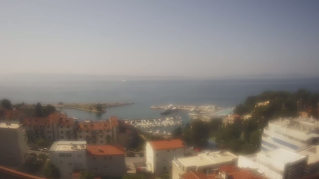 A hazy, sunny day overlooks a calm sea, a marina filled with boats, and a collection of buildings nestled on a hillside.