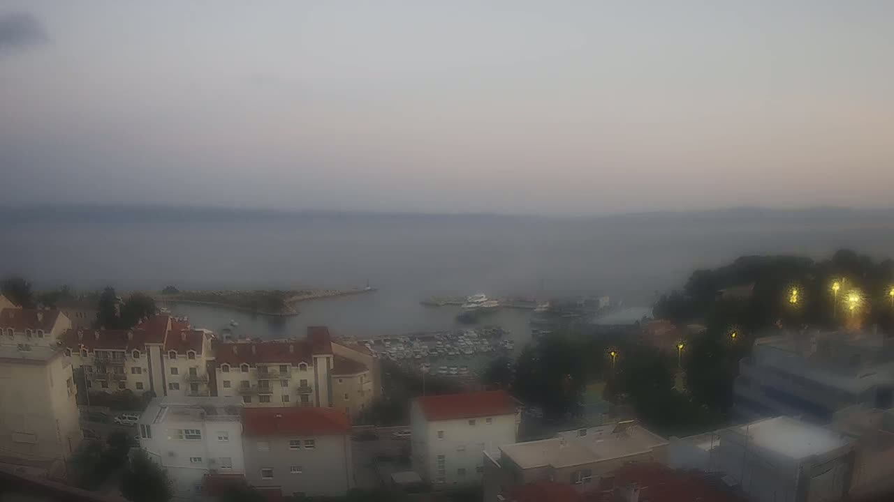 A hazy, dusk view overlooks a small harbor filled with boats, adjacent to a town with low-rise buildings.