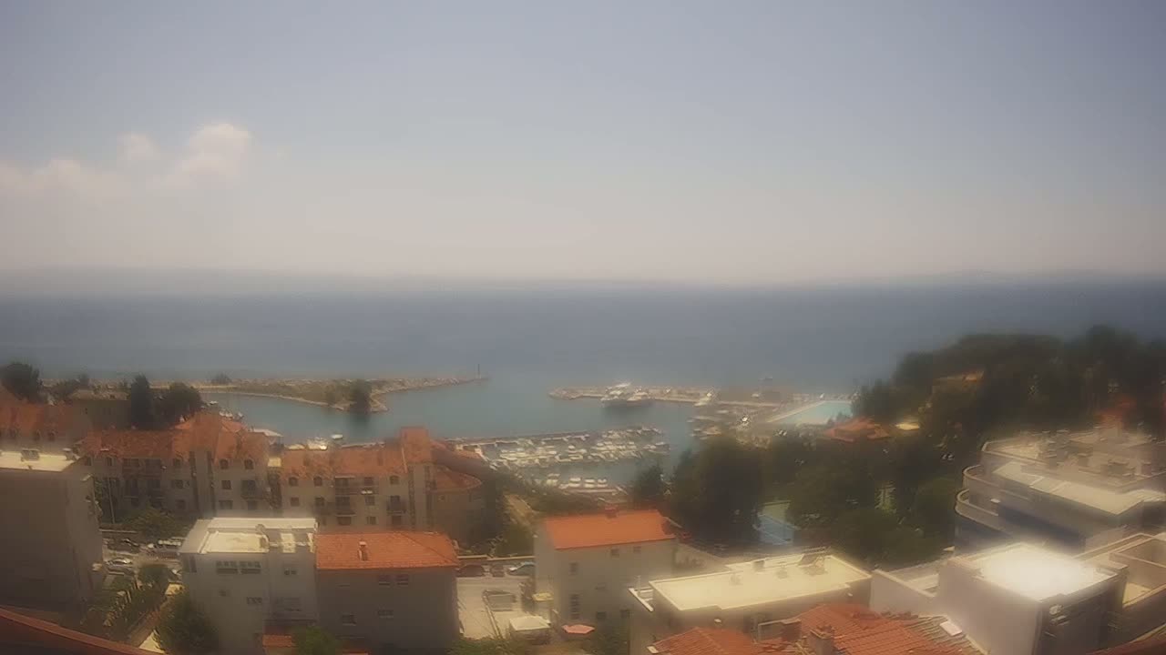 A hazy, sunny day overlooks a coastal town with a marina filled with boats, surrounded by buildings with red-tiled roofs and green vegetation.
