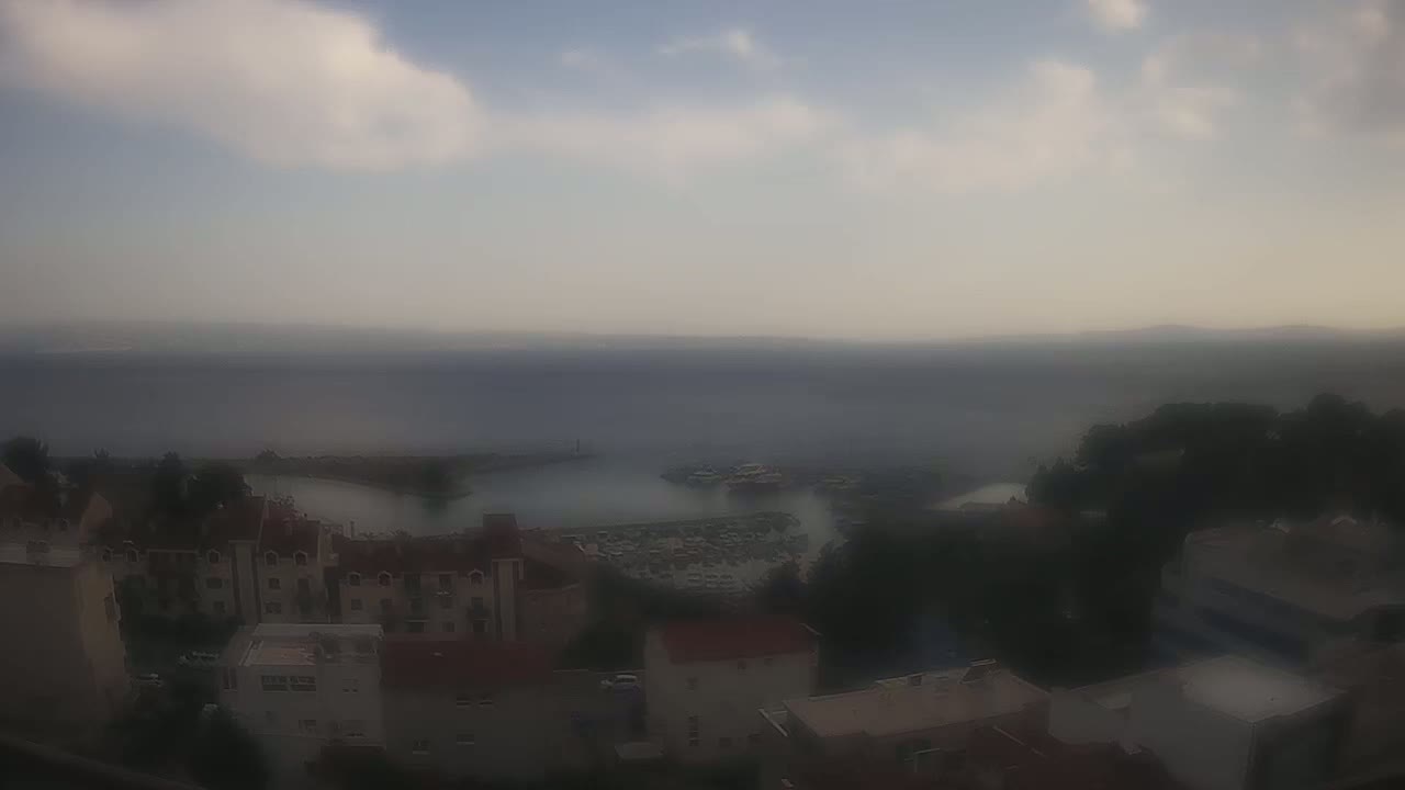 A hazy, overcast day reveals a coastal town with buildings clustered near a calm harbor, boats moored in the marina, and a distant, indistinct shoreline.