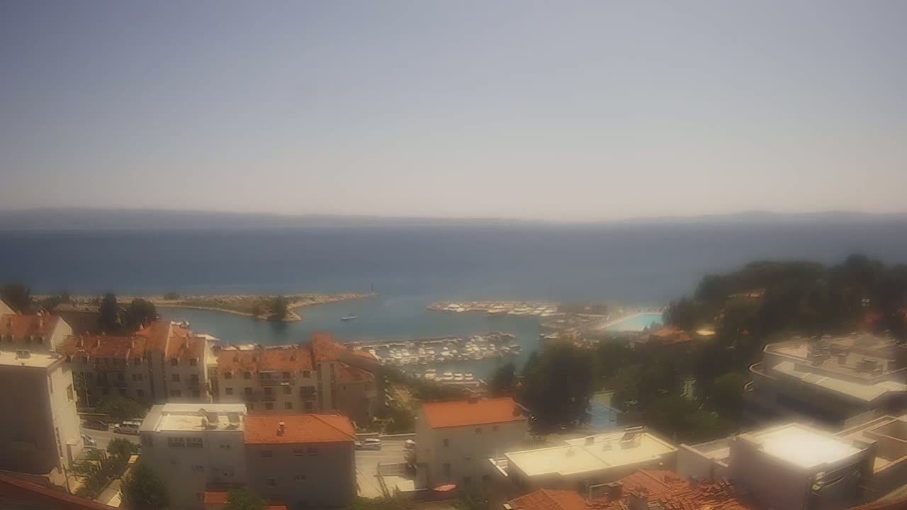 A hazy, sunny day overlooks a coastal town with red-roofed buildings clustered around a harbor filled with boats, all set against a calm sea and distant mountains.