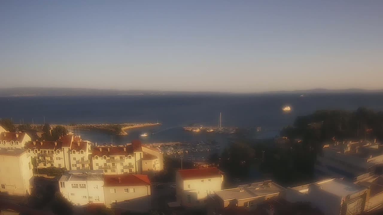 A coastal town with buildings and a marina is visible under a clear, sunny sky, overlooking a calm body of water.