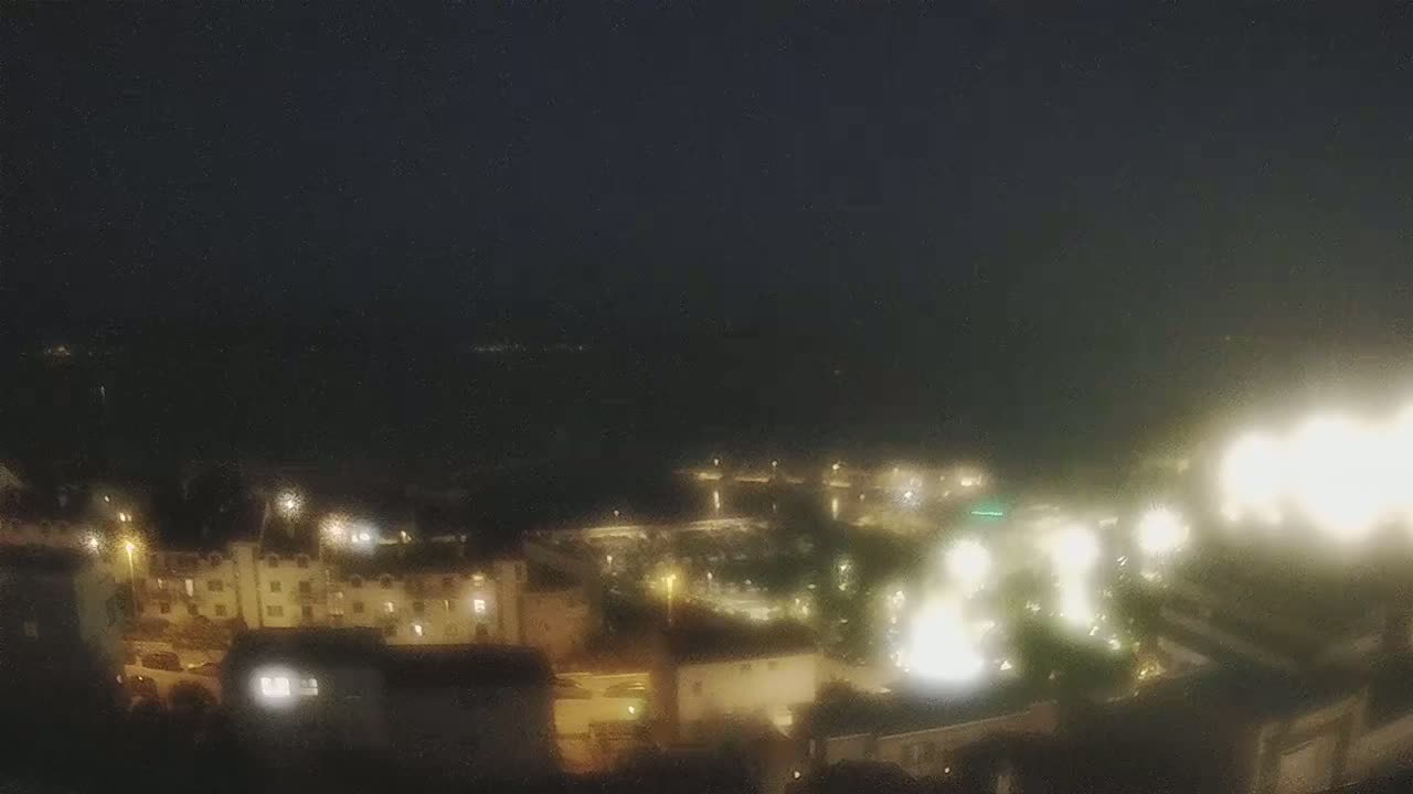 A nighttime, slightly hazy view shows a coastal town with buildings illuminated by lights, extending toward a dark body of water.