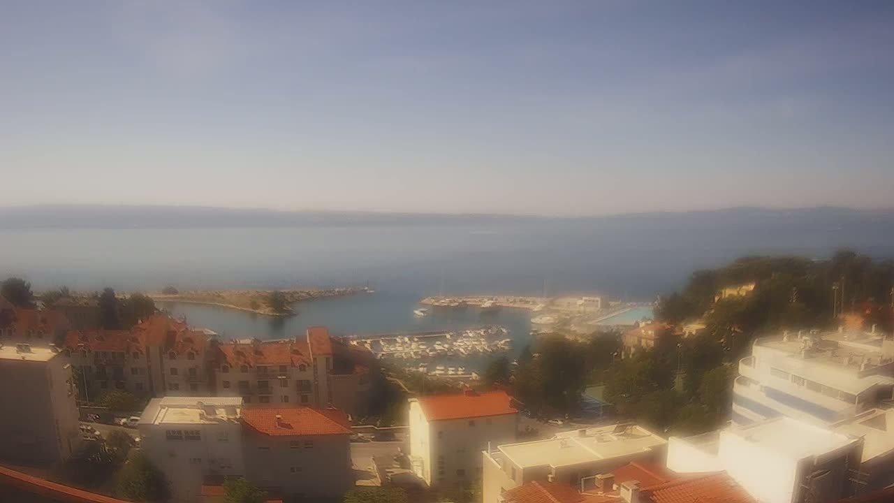 A hazy, sunny day overlooks a coastal town with a marina filled with boats,  buildings with red-tiled roofs, and green hillsides.