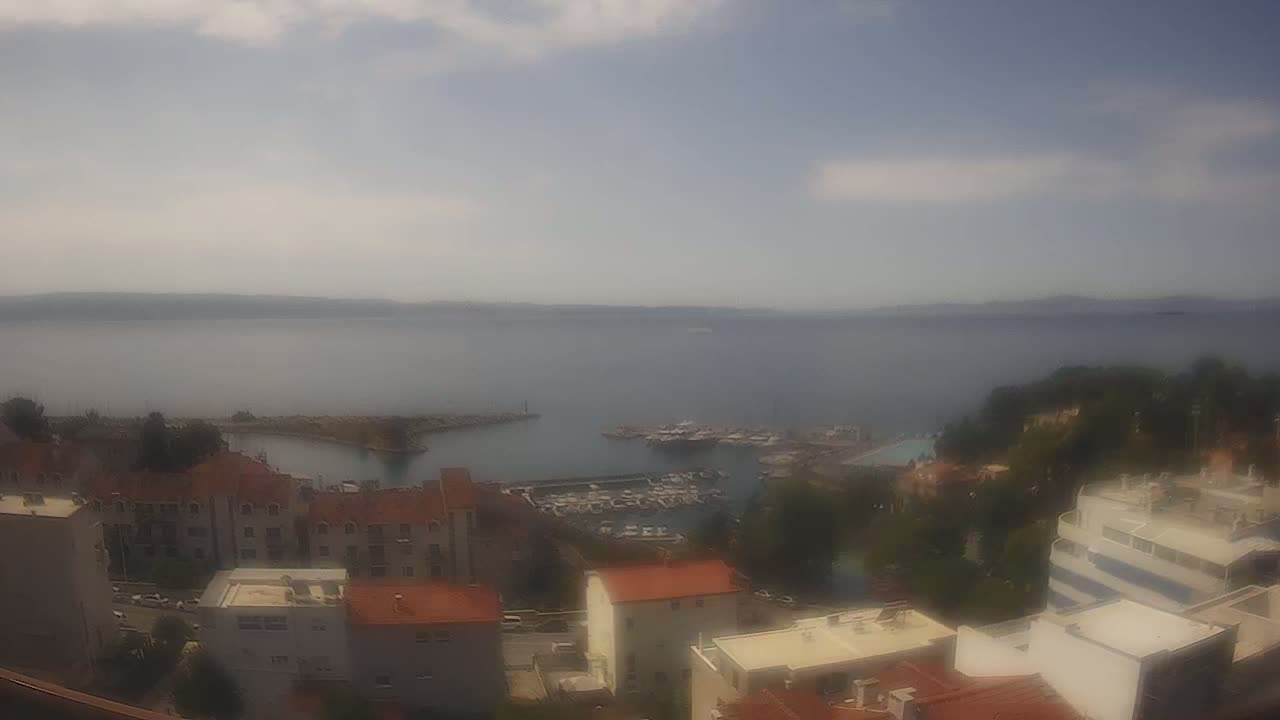 A hazy, partly cloudy day overlooks a coastal town with a marina filled with boats, and the ocean stretches to distant hills.