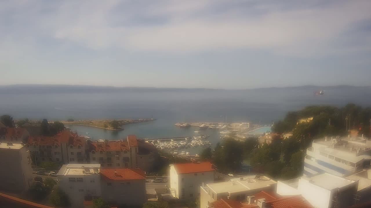 A hazy view of a coastal town with a marina filled with boats, under a mostly cloudy sky.