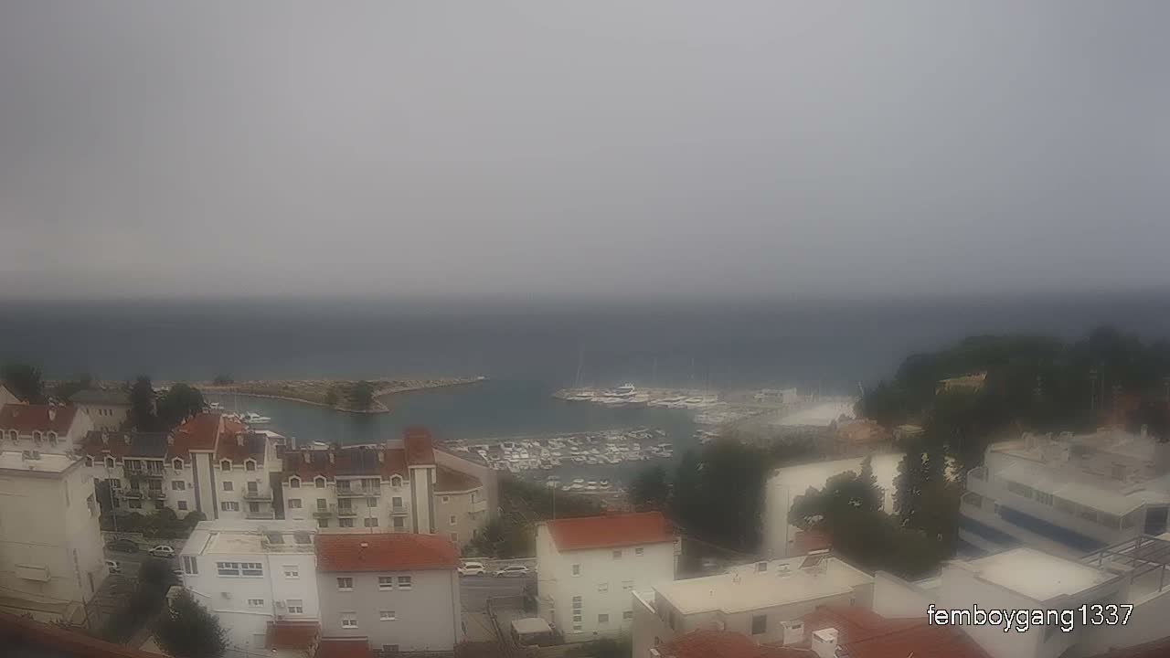 A hazy, sunny day overlooks a coastal town with red-roofed buildings surrounding a harbor filled with boats.