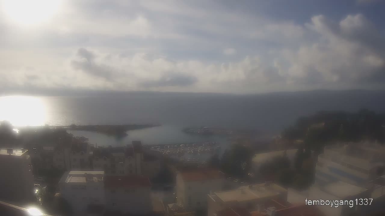 An elevated view reveals a coastal town with buildings and a marina filled with boats along a bay, all under a bright, partly cloudy sky with significant sun glare reflecting on the water.
