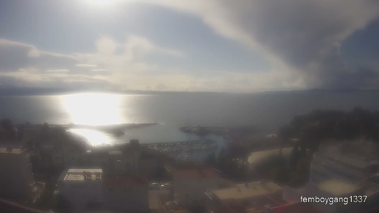 An elevated, somewhat hazy view on a partly cloudy day reveals bright sunlight reflecting off a large body of water, highlighting a harbor filled with boats and surrounding coastal buildings.