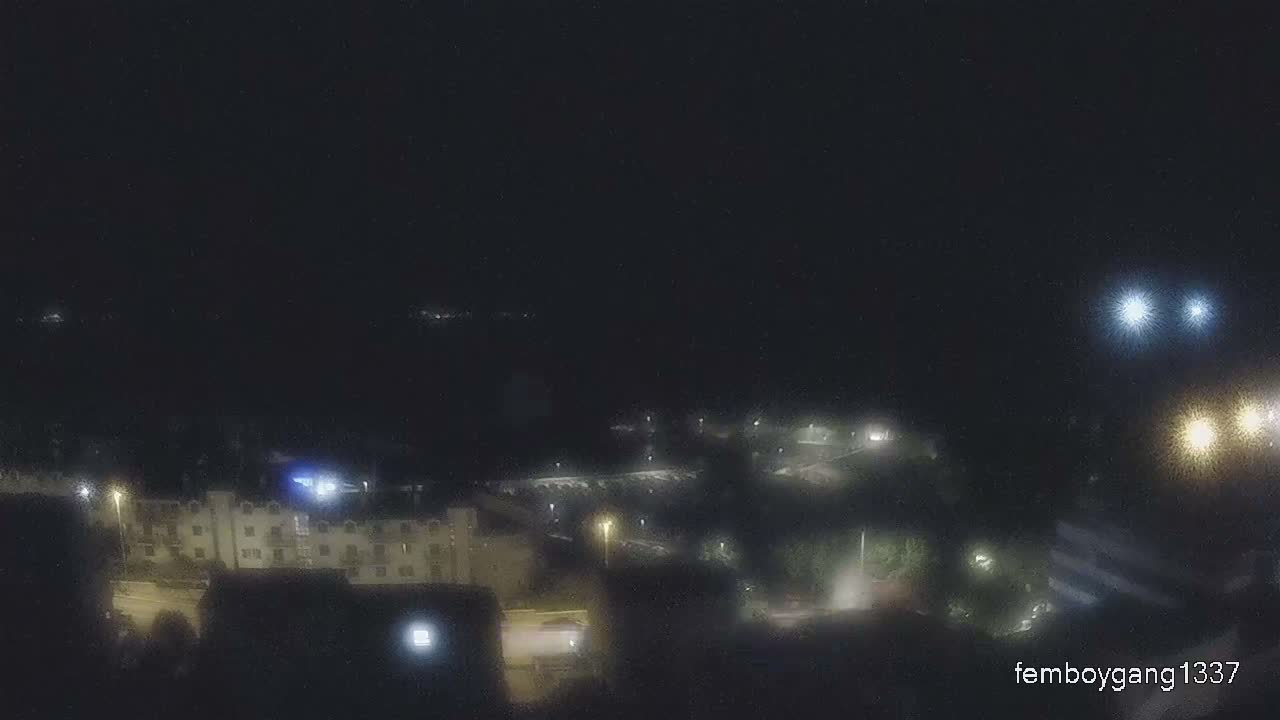 A dark, blurry nighttime view of a town or city features illuminated buildings, streetlights, and distant bright lights against a clear, dark sky.