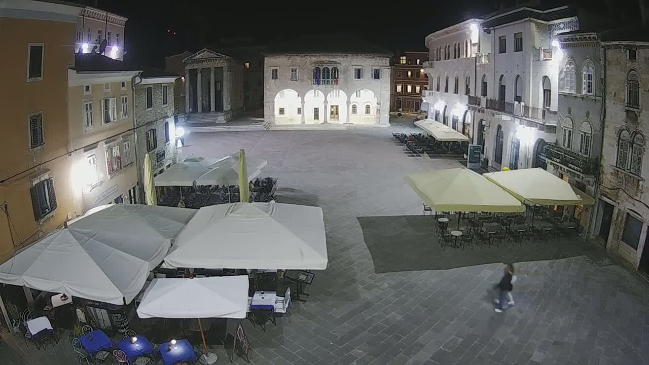 A nighttime view of a town square with several outdoor café umbrellas set up, surrounded by buildings.