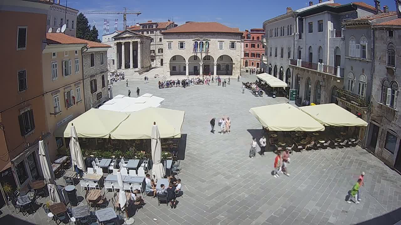 A large town square with several outdoor cafes under awning-covered seating, surrounded by historic buildings on a sunny day.