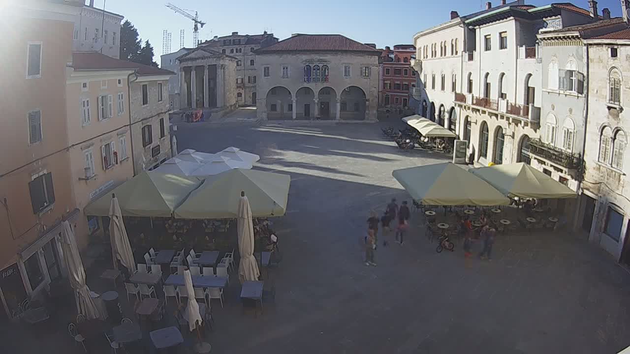 A sunny, high-angle view of a town square with several outdoor cafes under umbrellas, surrounded by old buildings.