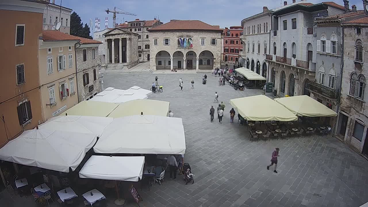 A town square with several outdoor cafes under umbrellas, surrounded by historical buildings on a partly cloudy day.