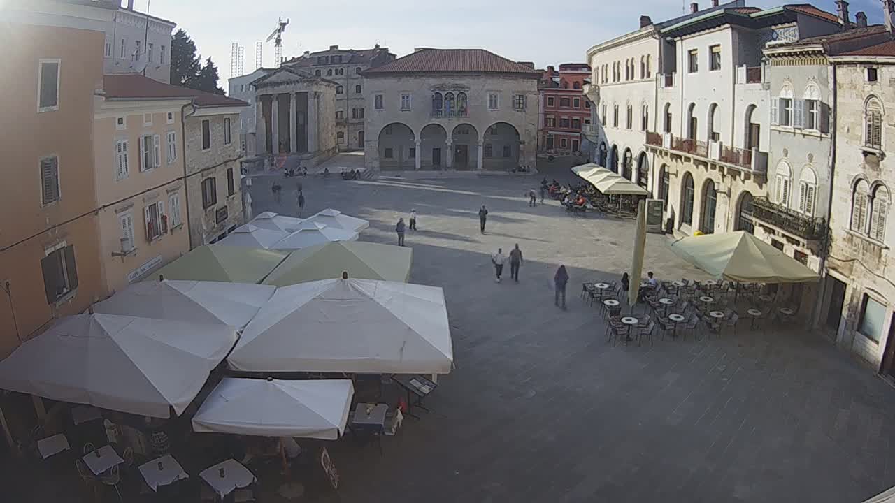 A town square with several outdoor cafes under umbrellas, surrounded by old buildings, on a partly sunny day.