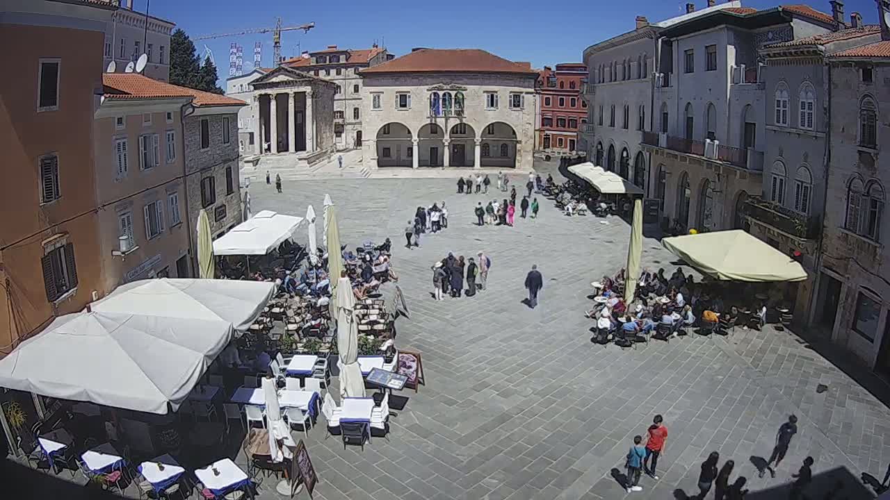 A sunny town square with several outdoor cafes and people sitting at tables, surrounded by historical buildings.