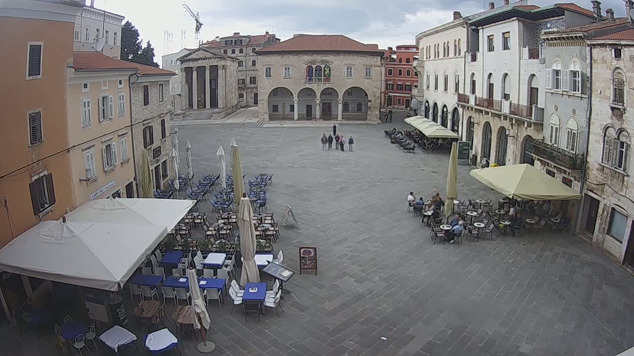 A large town square with several outdoor cafes under umbrellas, surrounded by historic buildings, on an overcast day.