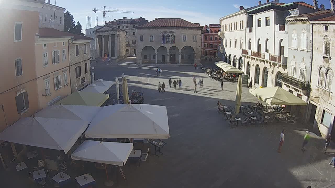 A sunny day reveals a town square with several people walking and sitting at outdoor cafes, surrounded by historic buildings.