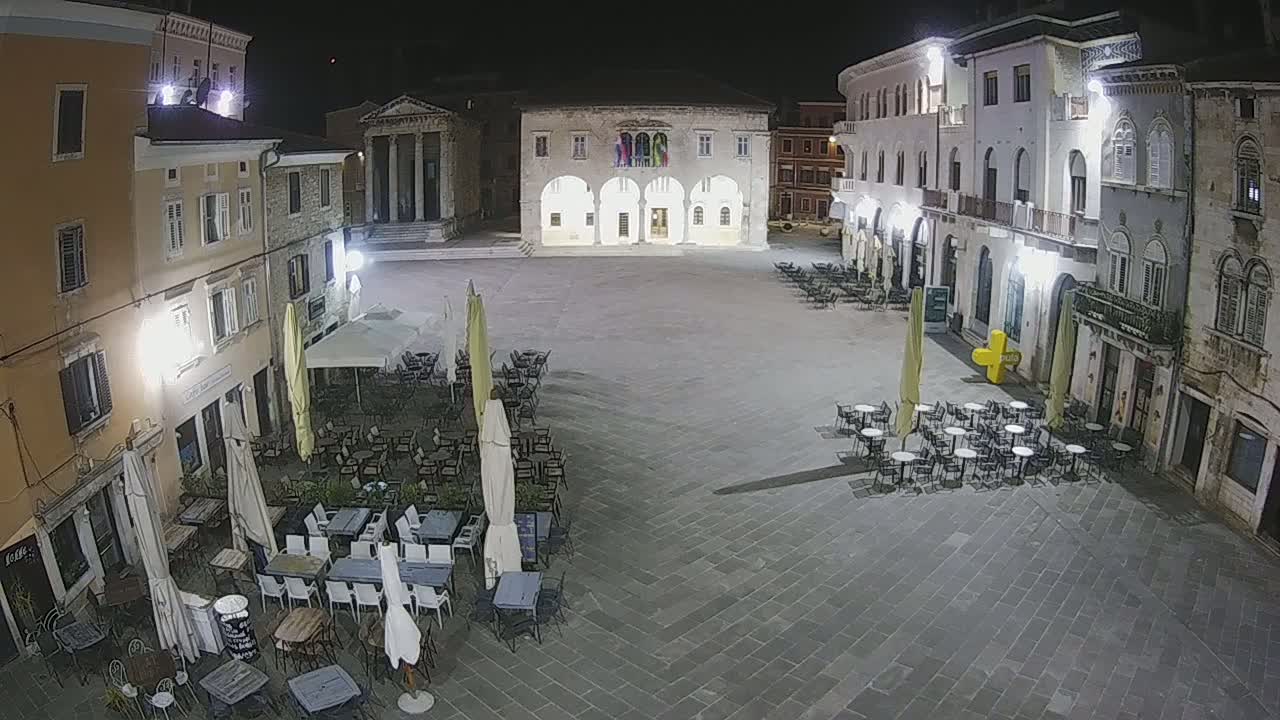 A nighttime, outdoor view of a town square with several buildings surrounding it, multiple café tables and chairs set up in the square under outdoor umbrellas.