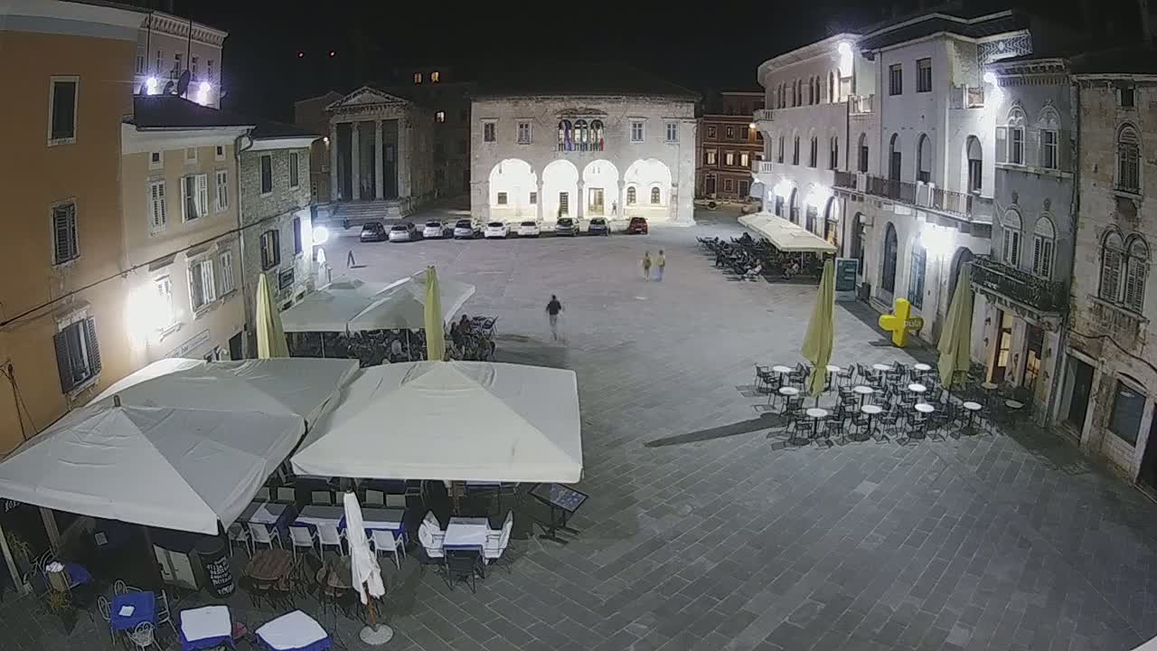 A nighttime view of a town square with several outdoor cafes under umbrellas, surrounded by stone buildings under artificial light.