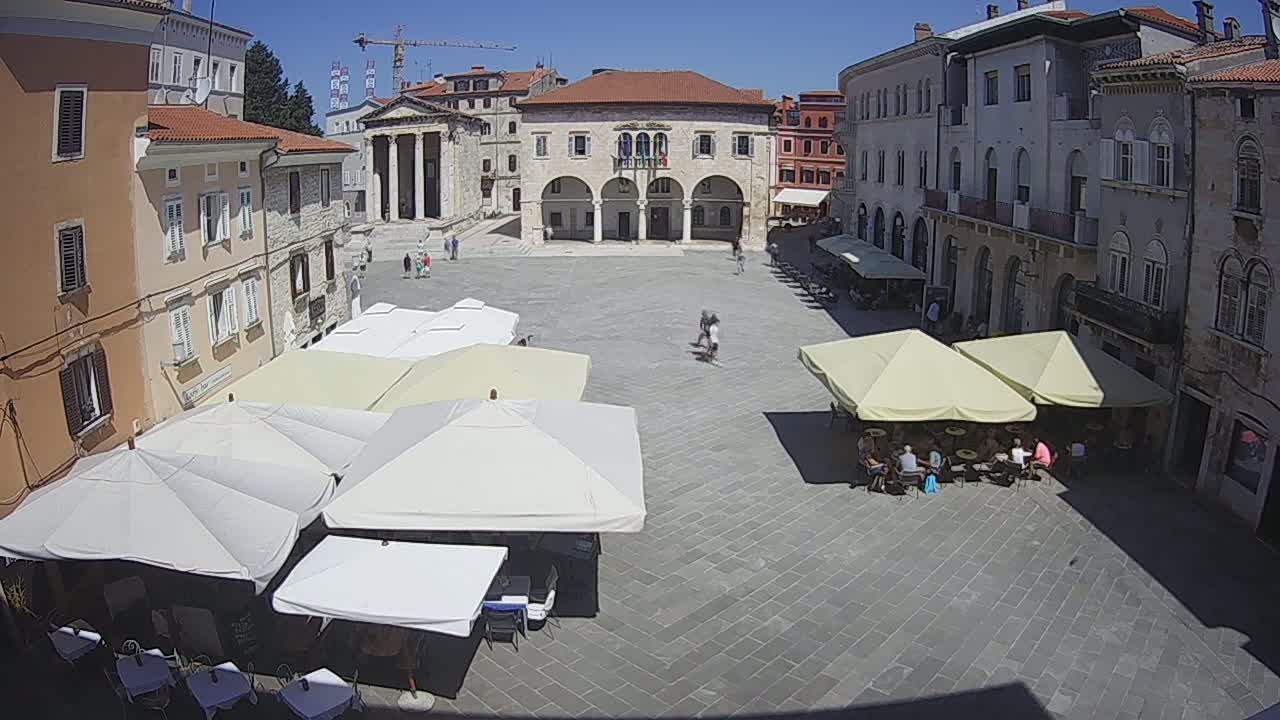 A sun-drenched town square, filled with outdoor cafe umbrellas and a few people, is surrounded by historic buildings.