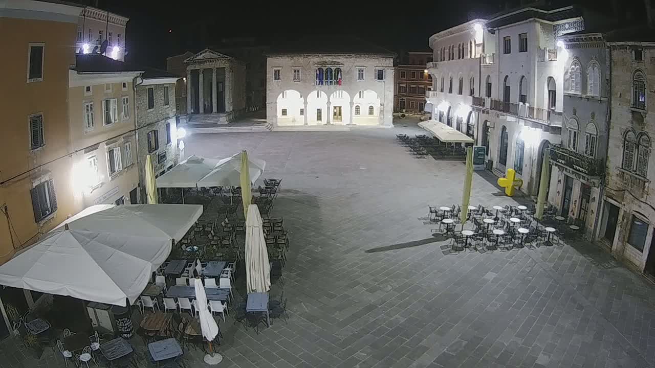 At night, a town square with several outdoor cafes under umbrellas is illuminated, surrounded by stone buildings.