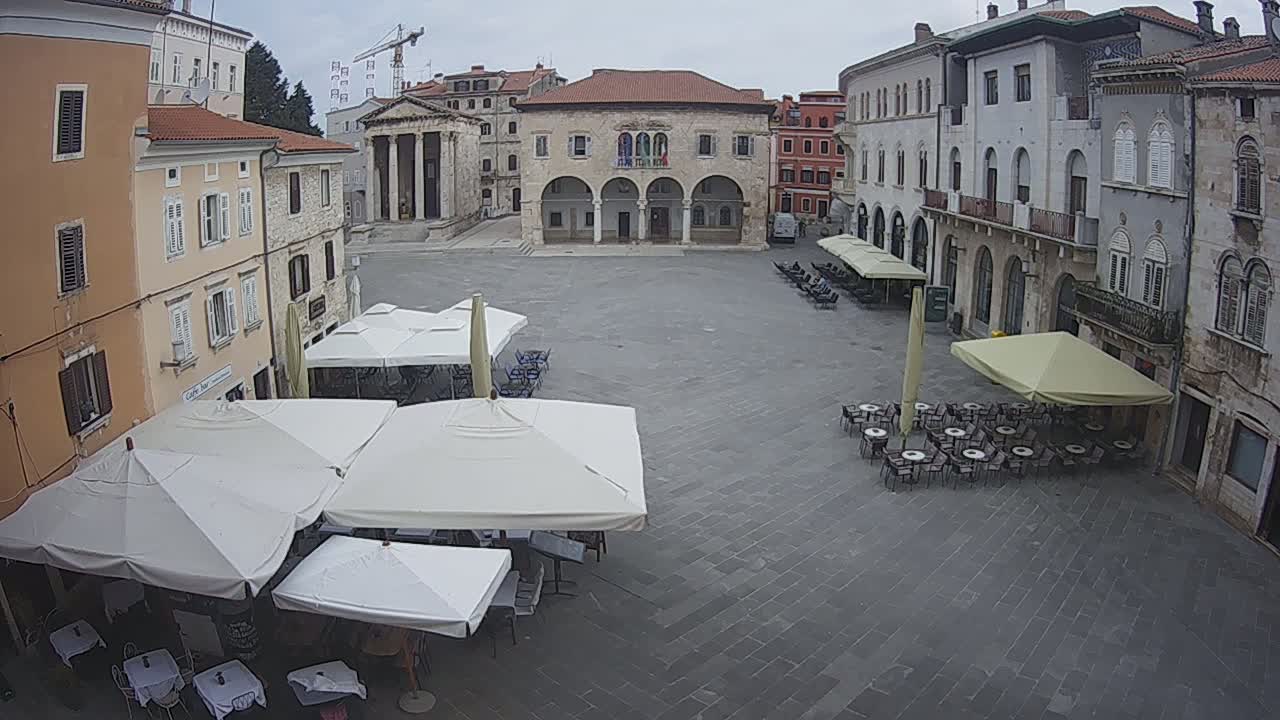 An overcast day in a European town square with several outdoor cafe umbrellas and tables set up in front of historic stone buildings.