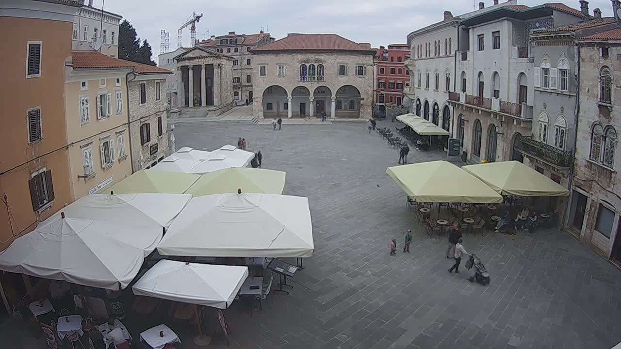 An overcast day in a European town square with several outdoor cafe umbrellas and stone buildings surrounding a large paved plaza.