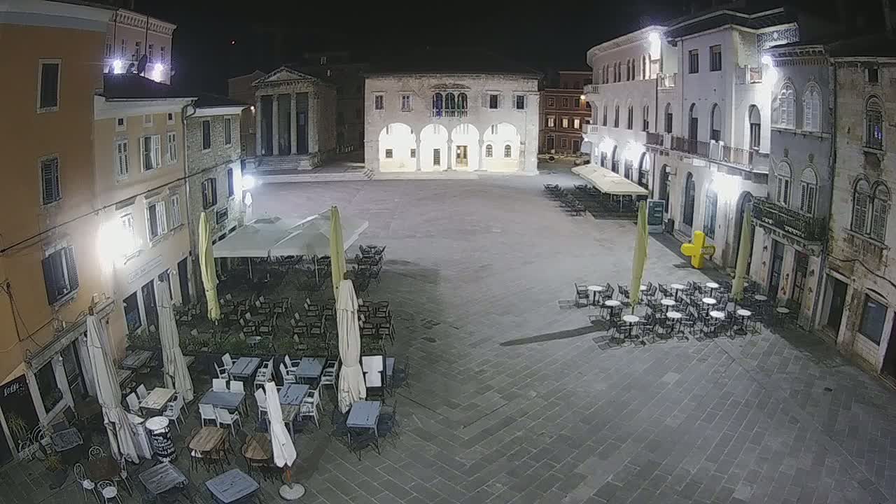 A nighttime view of a town square with several outdoor cafe seating areas, surrounded by stone buildings.